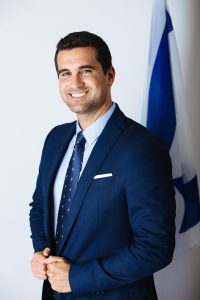 Smiling man in a suit stands confidently with hands clasped, a flag in the background, representing leadership and professionalism.